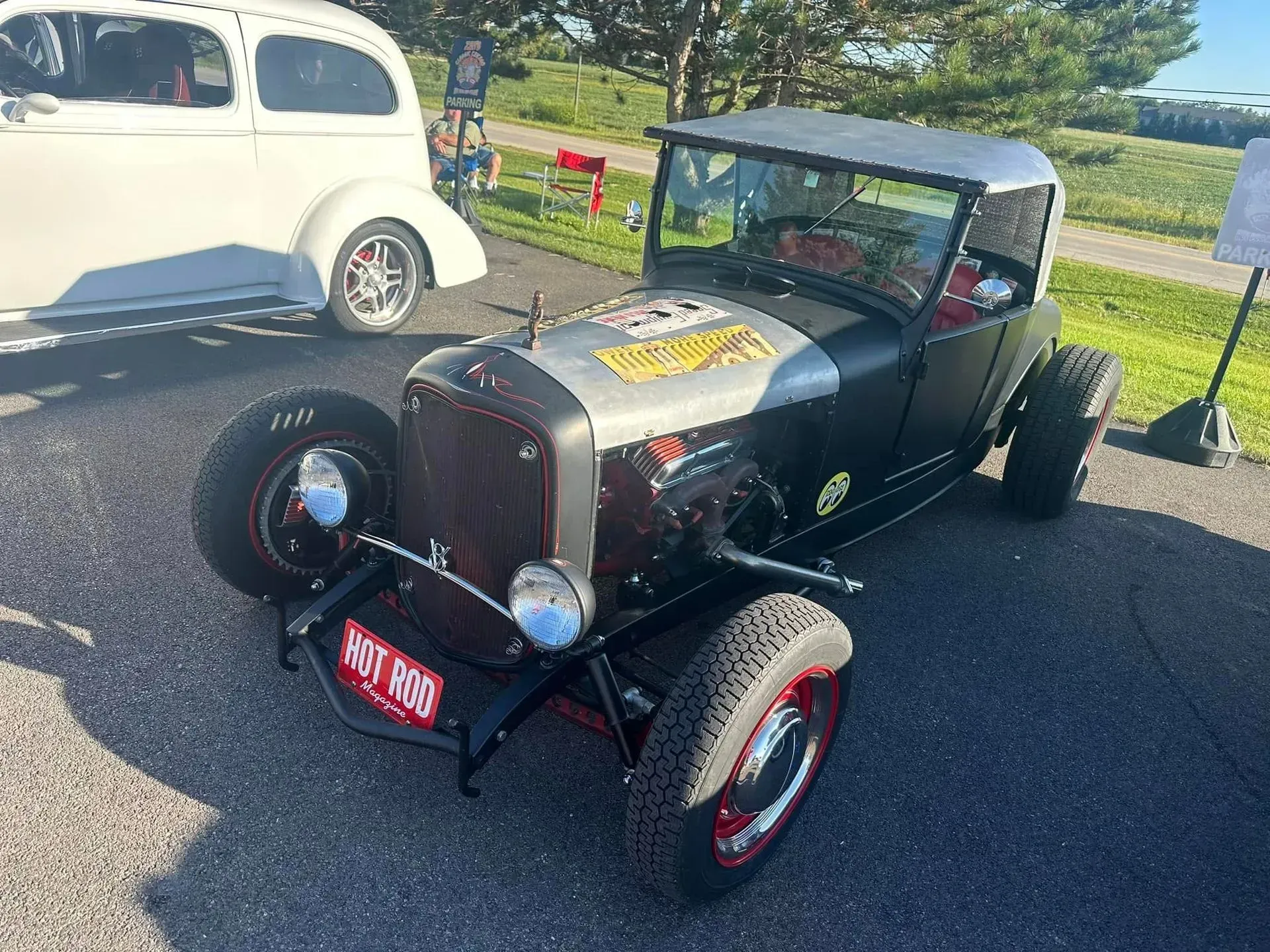 A black and silver vintage hot rod car parked on an asphalt lot near a white classic car.