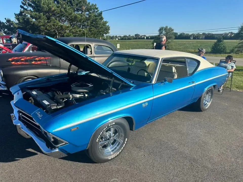A blue Chevrolet Chevelle SS with a white vinyl roof and open hood parked at a sunny outdoor car show.
