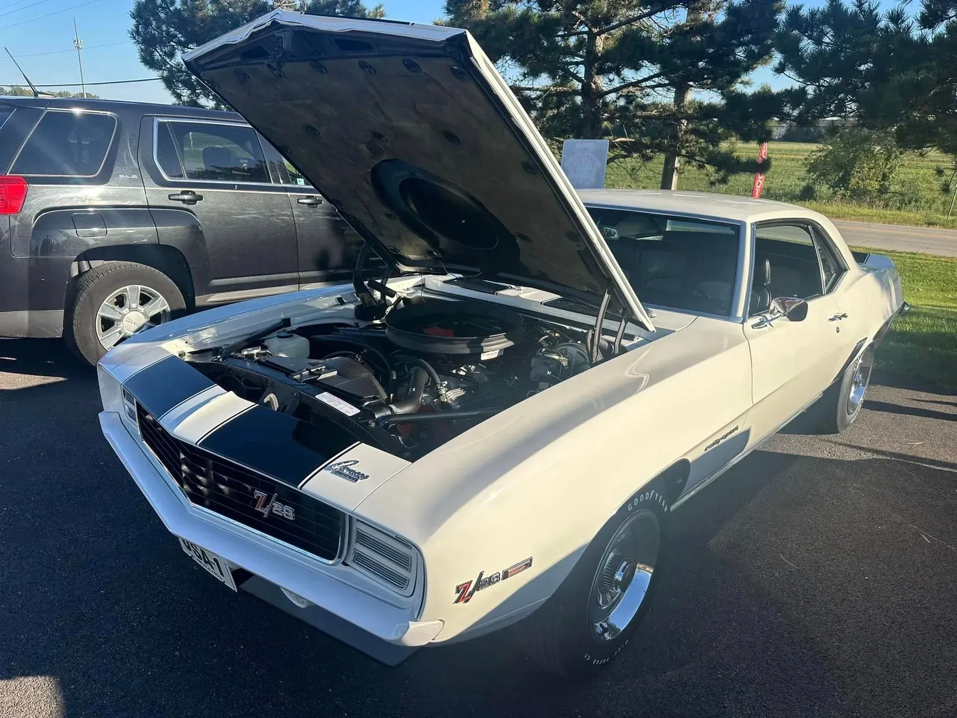 A white 1969 Chevrolet Camaro Z/28 with a black hood stripe and the hood open, parked on asphalt near a dark SUV.