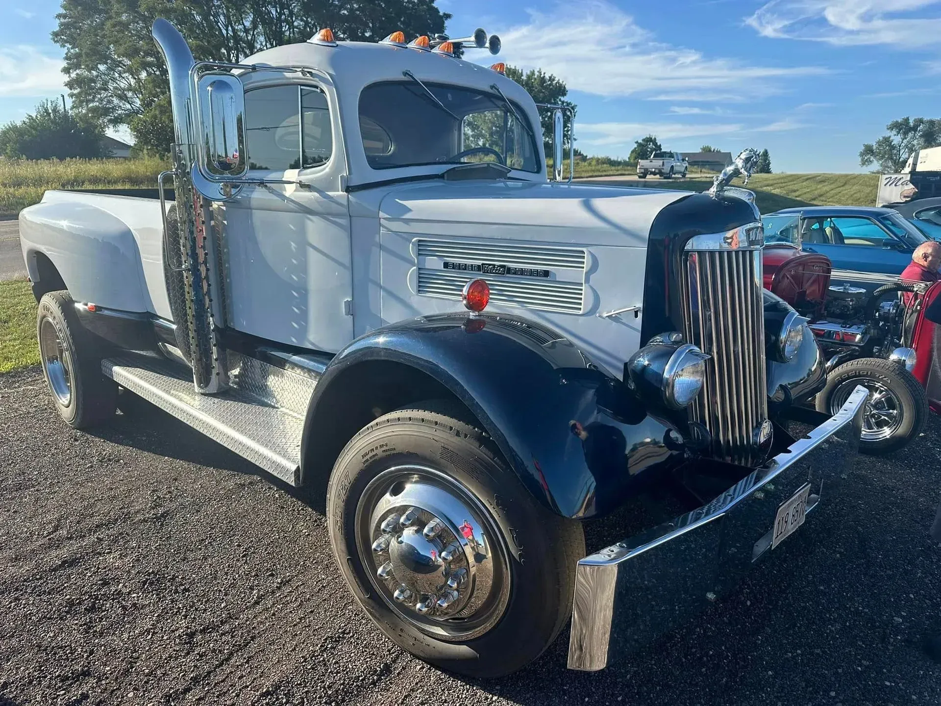 A white and black vintage-style custom pickup truck with a large chrome grille and exhaust stack parked on gravel.