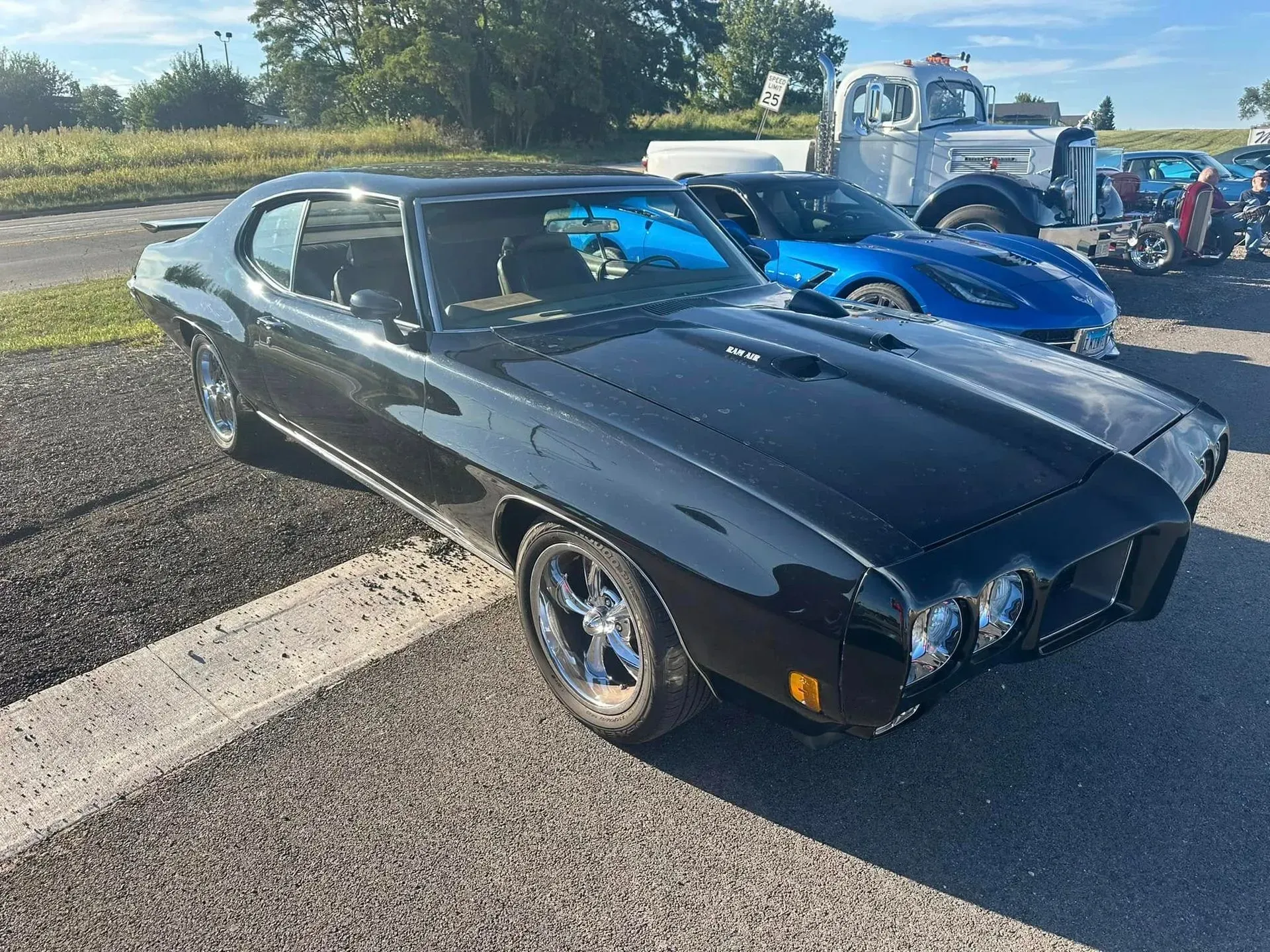 A shiny black classic Pontiac muscle car parked on an asphalt lot, with a blue sports car and a tow truck in the back.