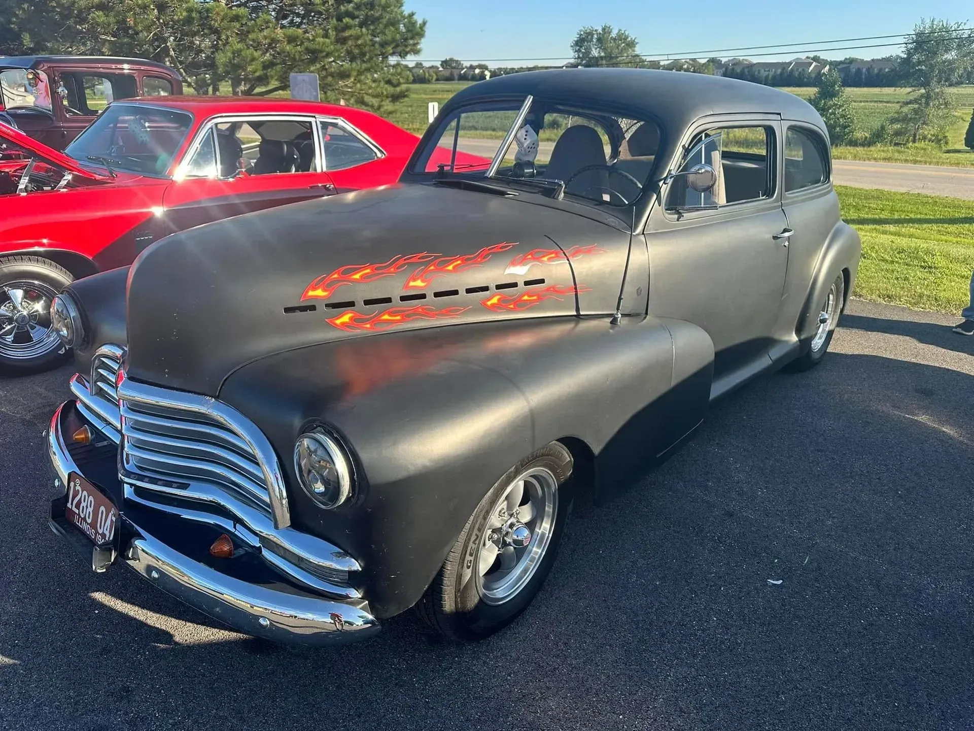 A matte black classic car with chrome accents and flame details parked on asphalt beside a bright red vehicle.