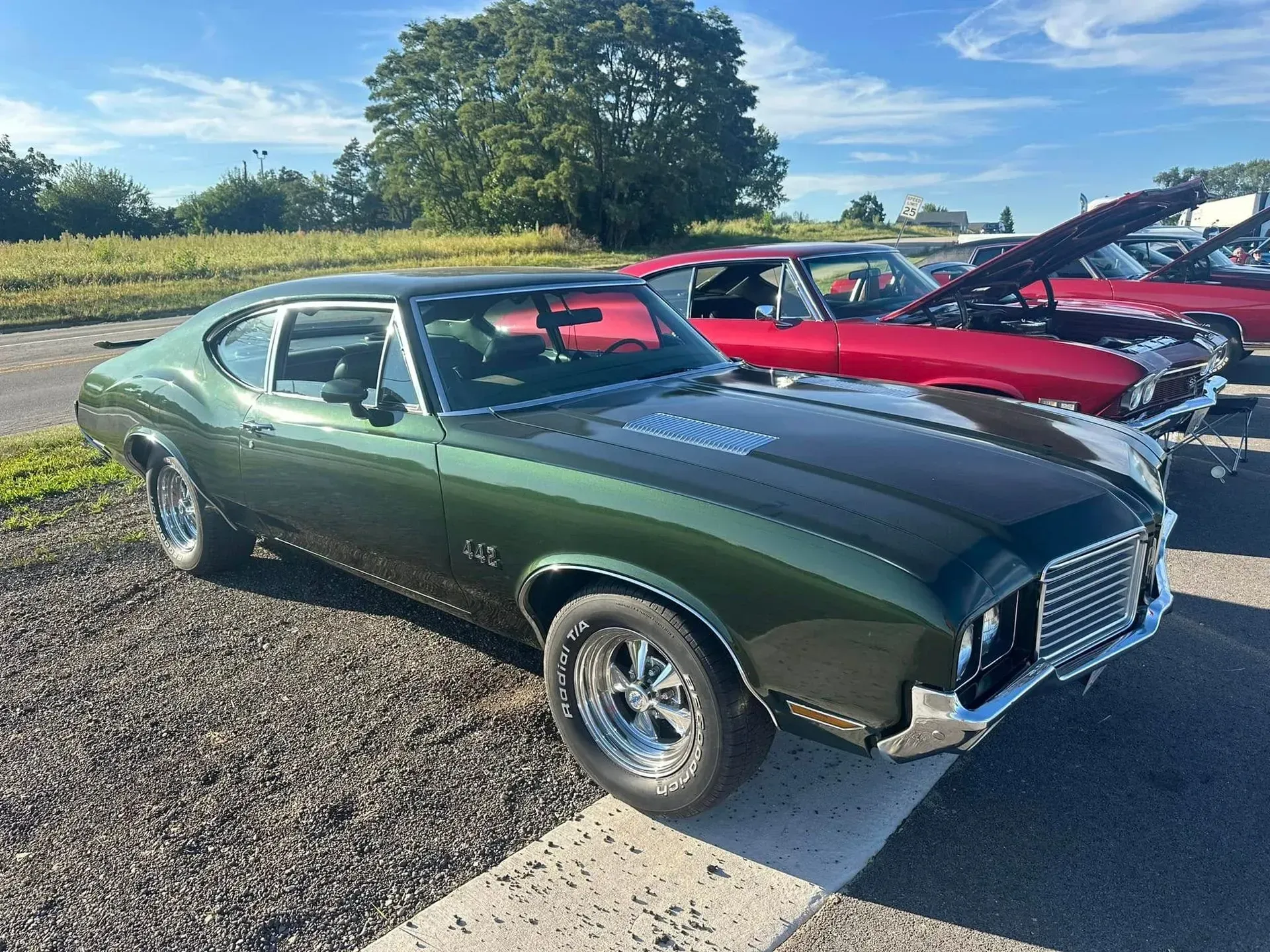 A dark green 1970 Oldsmobile 442 parked on asphalt next to a red car at an outdoor car show.