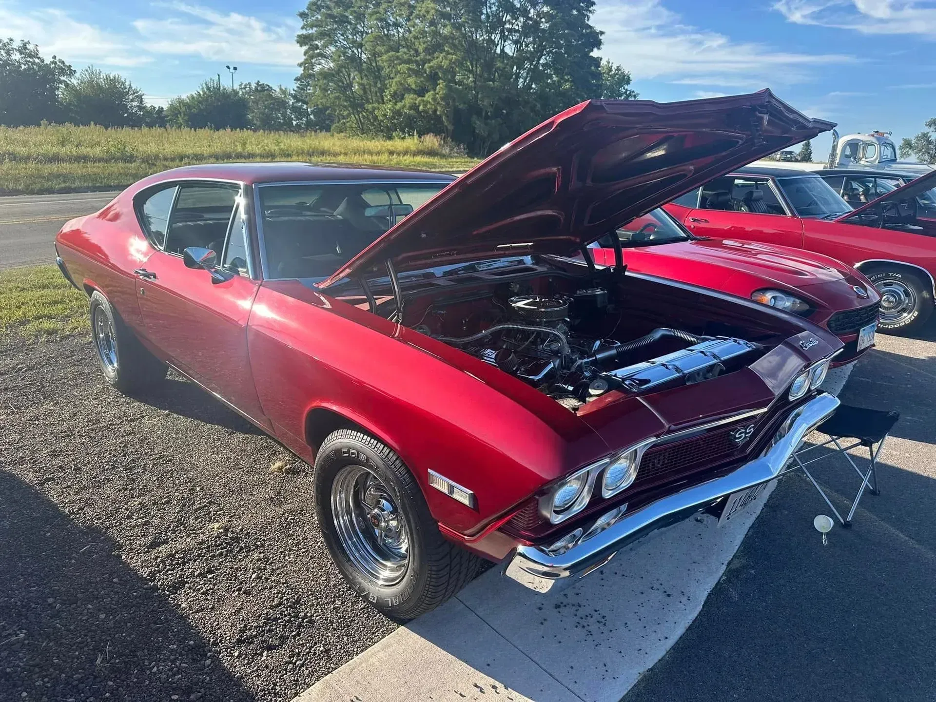 A shiny red classic muscle car with its hood open, parked on a gravel lot at an outdoor car show on a sunny day.