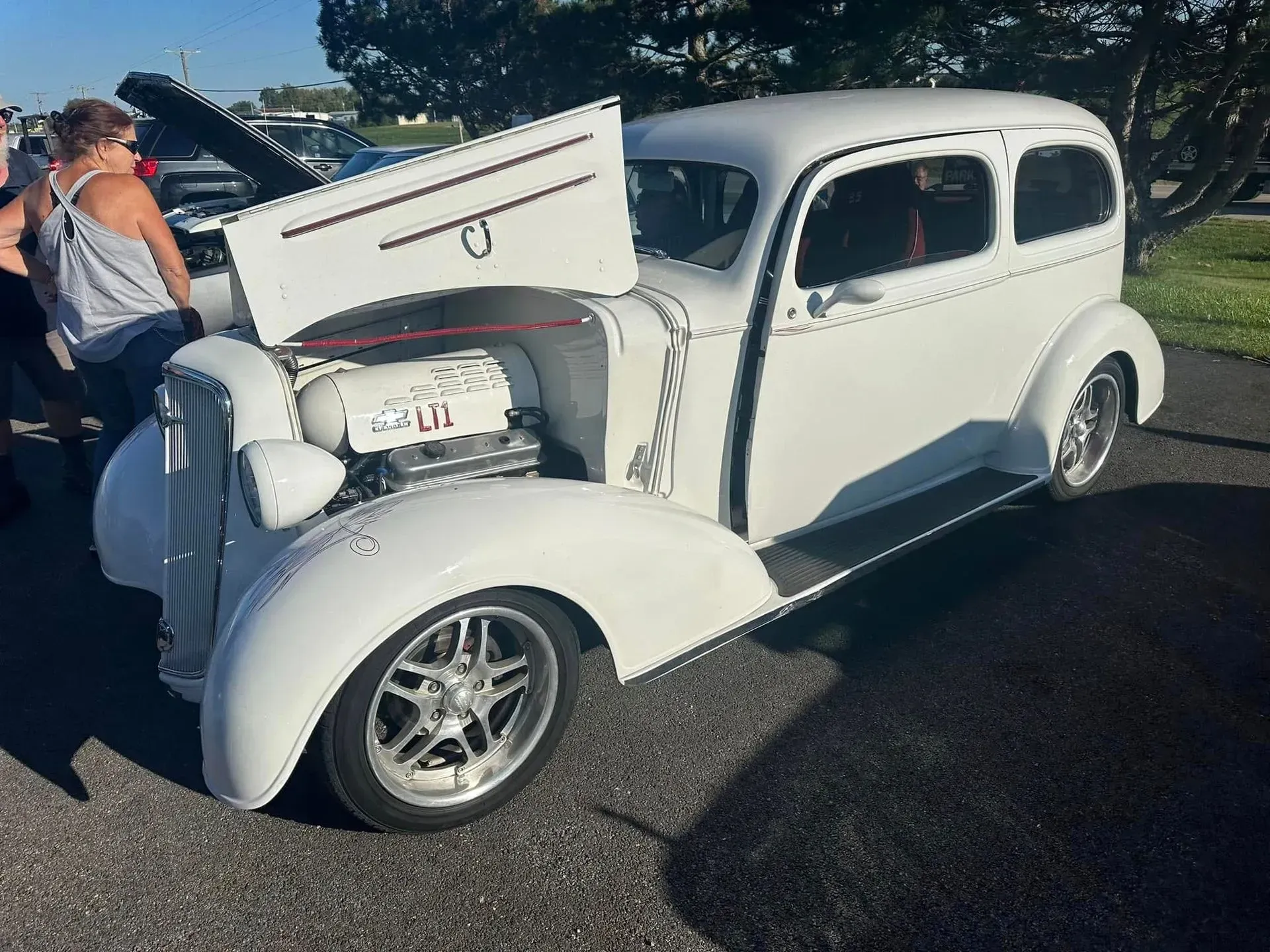 A white custom classic sedan with its hood open, parked on asphalt at an outdoor car show.