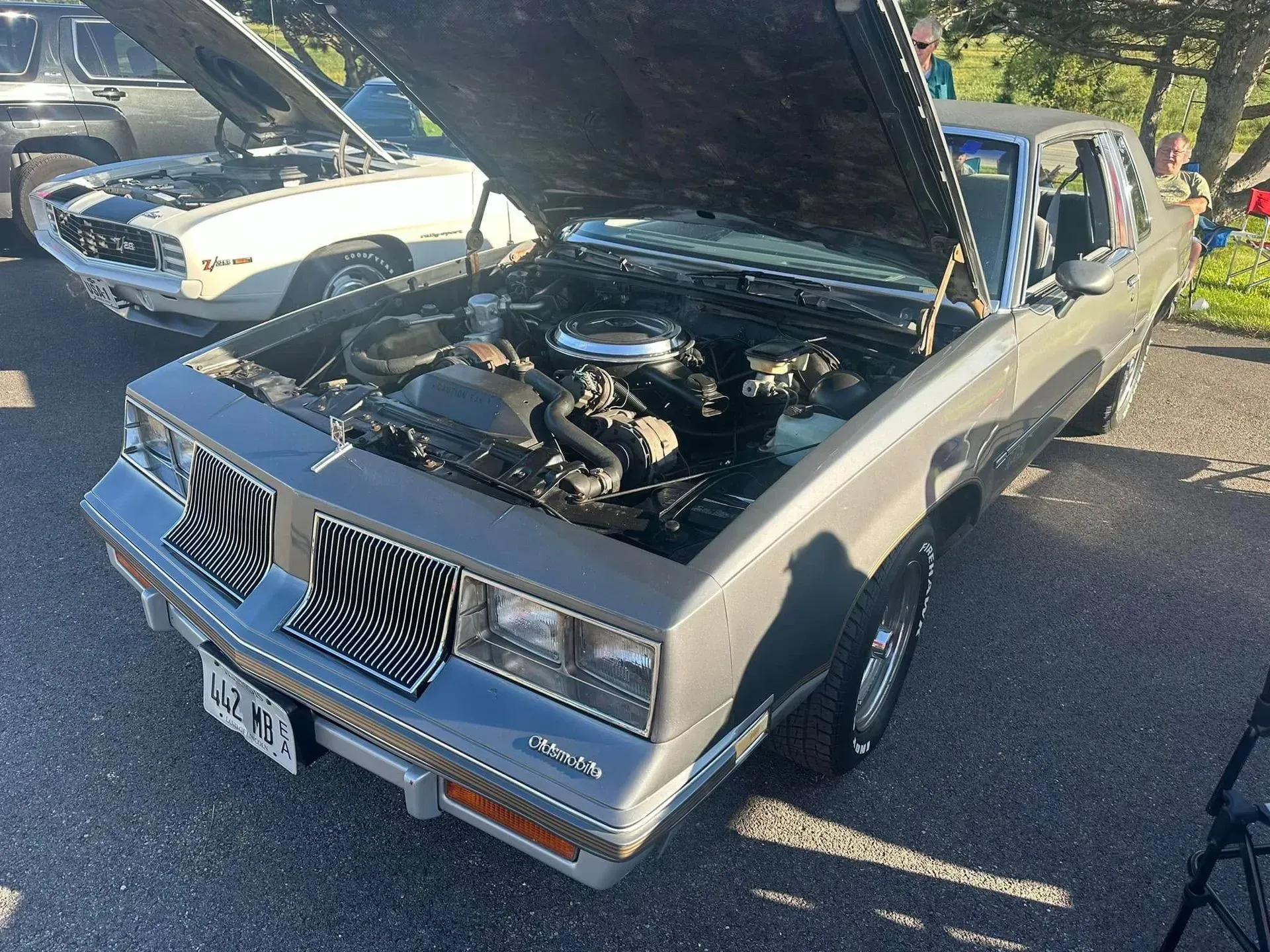 A grey Oldsmobile Cutlass parked with its hood open, showcasing the engine, next to a white car at an outdoor event.