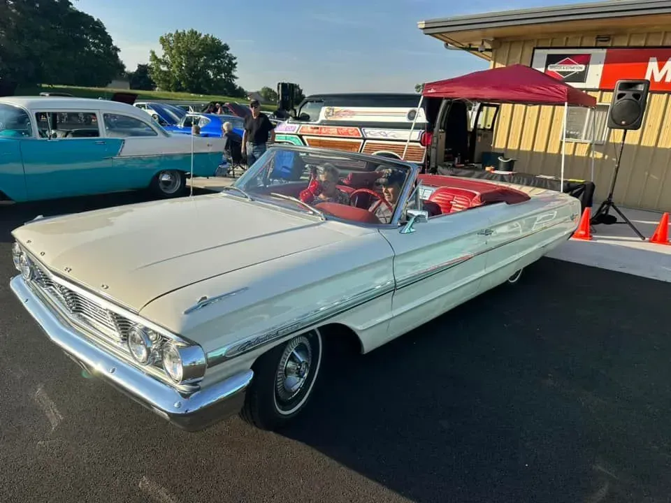 A white 1960s Ford convertible parked at an outdoor car show, with a red canopy and a blue vintage car in the background.