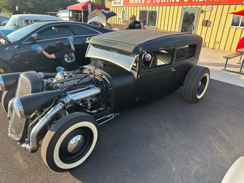 A matte black hot rod with white-wall tires parked outside a building as a person cleans a car in the background.