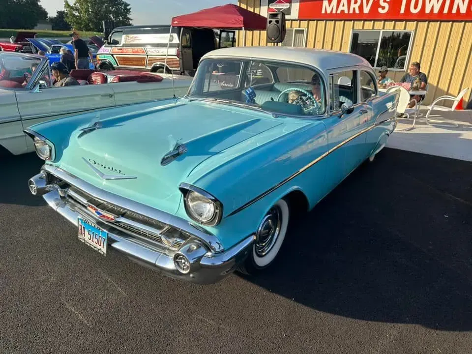 A vintage, two-tone blue and white 1957 Chevrolet parked outside a business building on a sunny day.