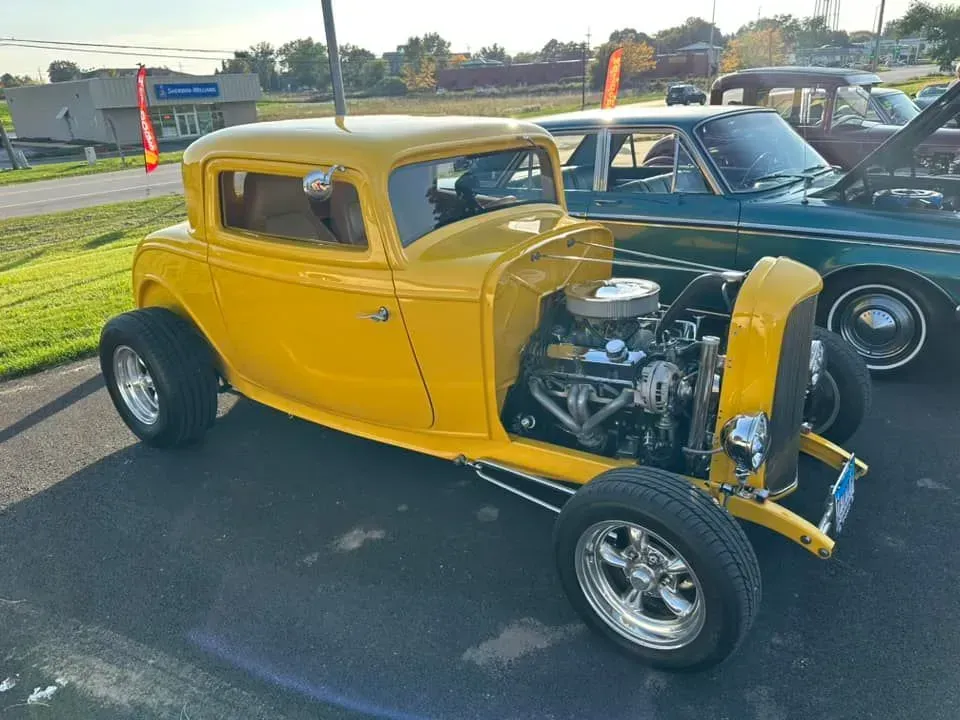 A bright yellow, vintage-style hot rod coupe parked on an asphalt lot with a dark green car in the background.