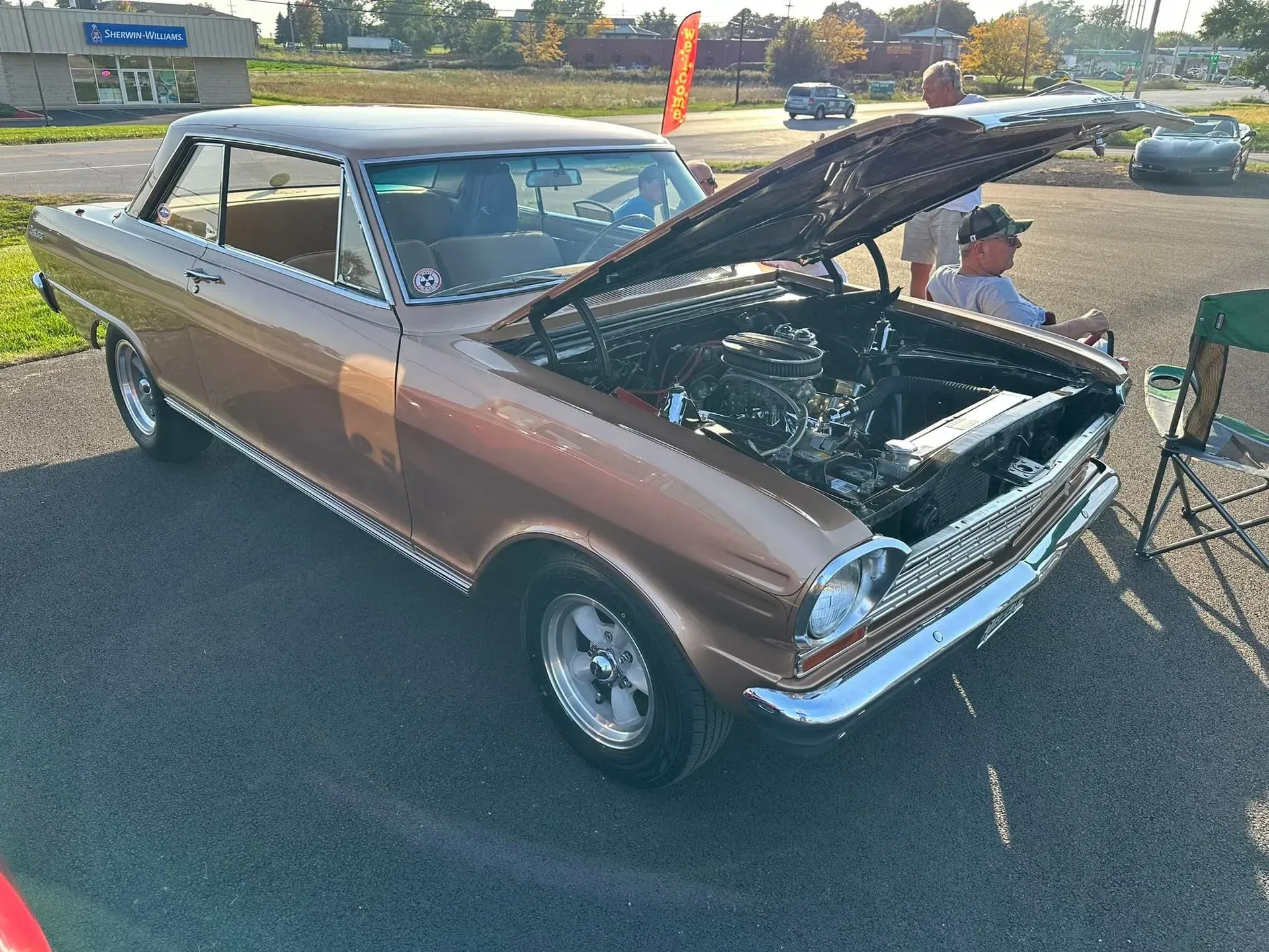 A metallic tan 1960s Chevy II Nova coupe parked with its hood open at an outdoor car show on a sunny day.