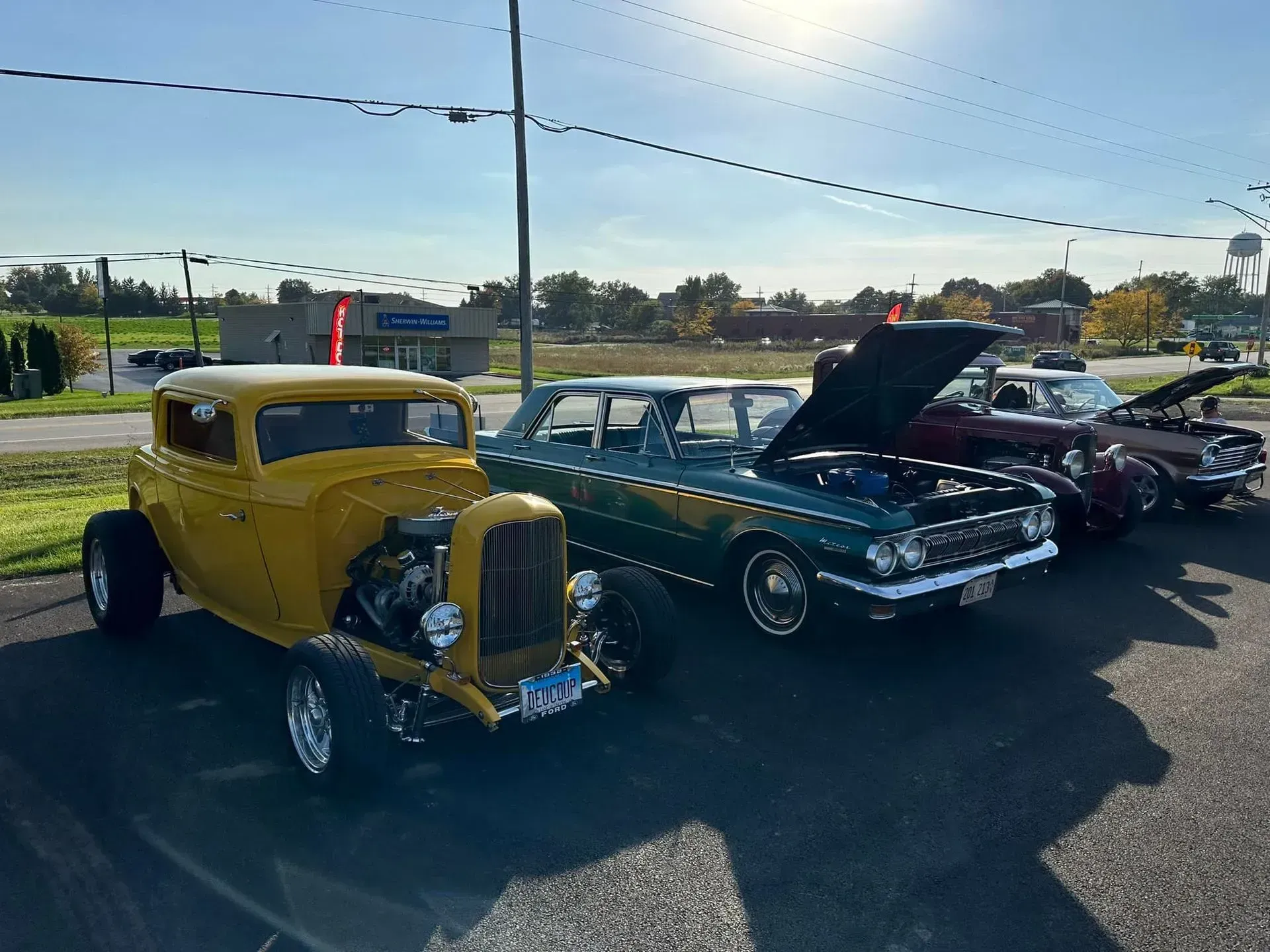 Three classic cars, including a yellow hot rod and a green vintage sedan, parked in an asphalt lot on a sunny day.