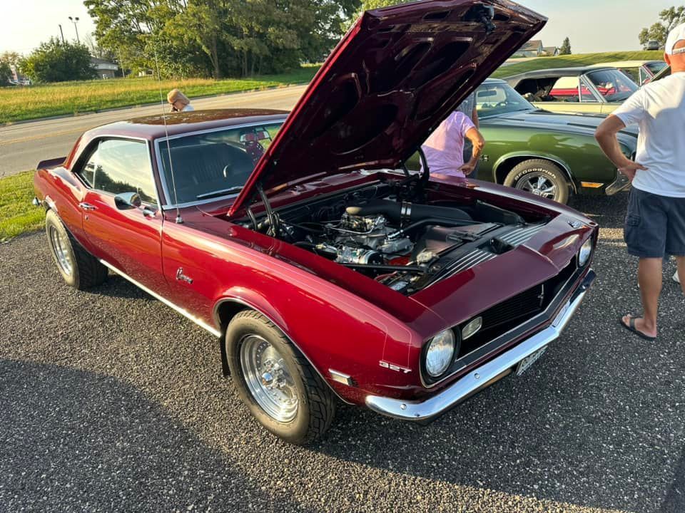 A dark red classic Chevrolet Camaro with its hood open, parked in a parking lot during a car show.