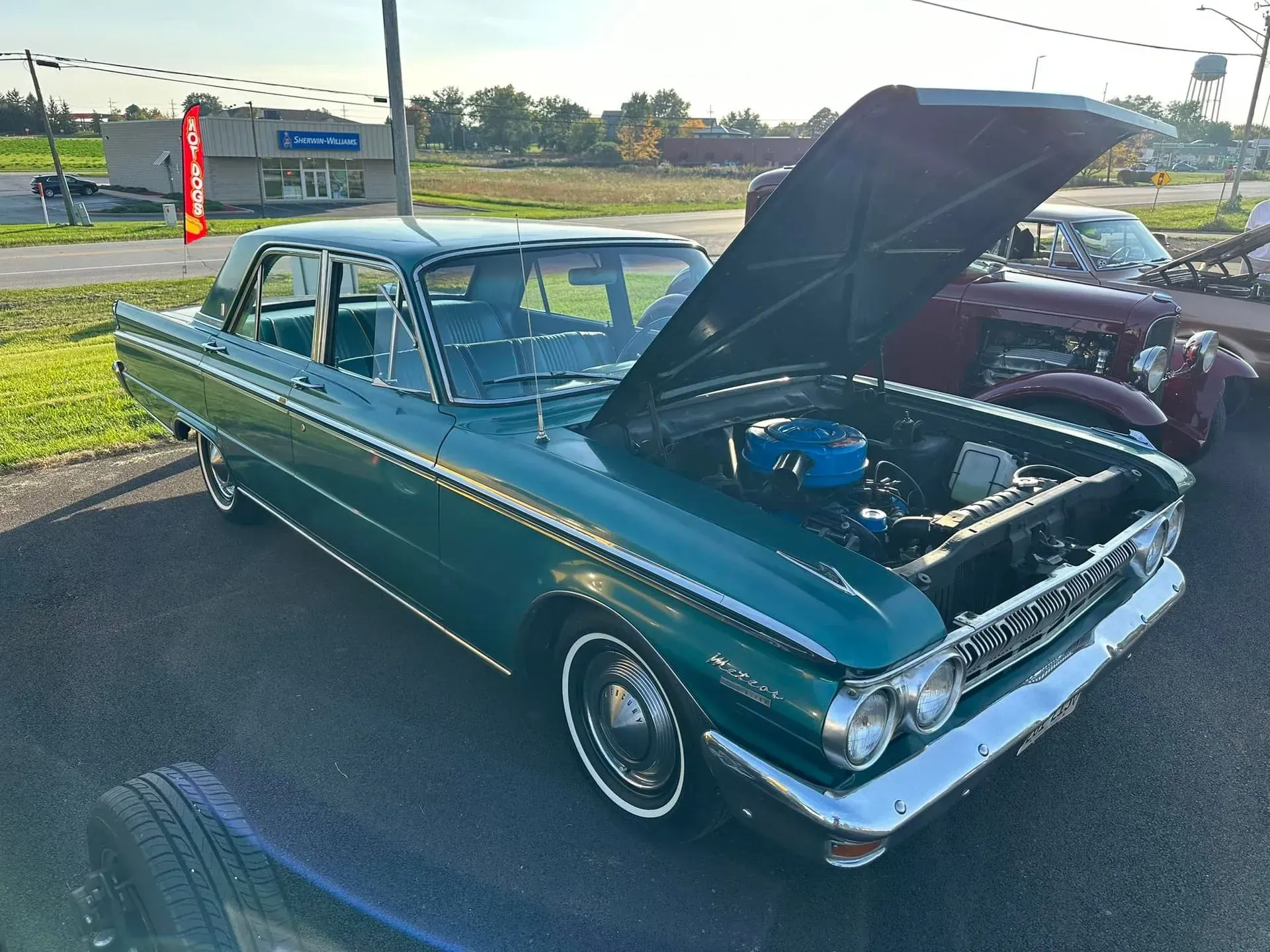 A dark green vintage sedan with its hood open, parked on asphalt in a parking lot during a sunny day.