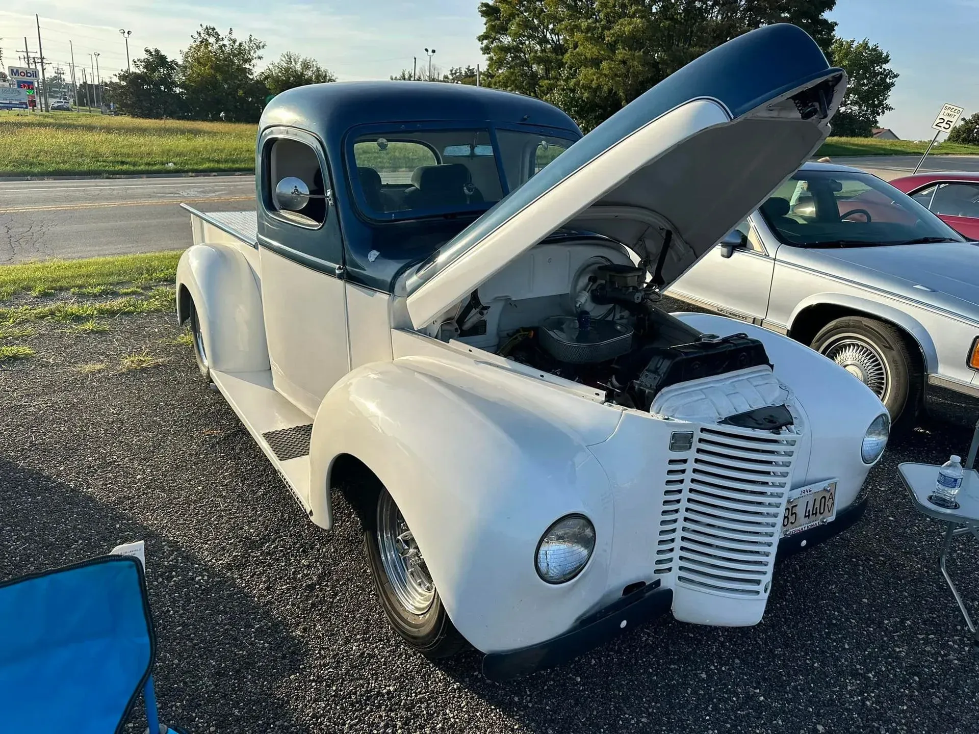 A vintage, two-tone blue and white truck with its hood open, parked on a gravel lot next to another car.