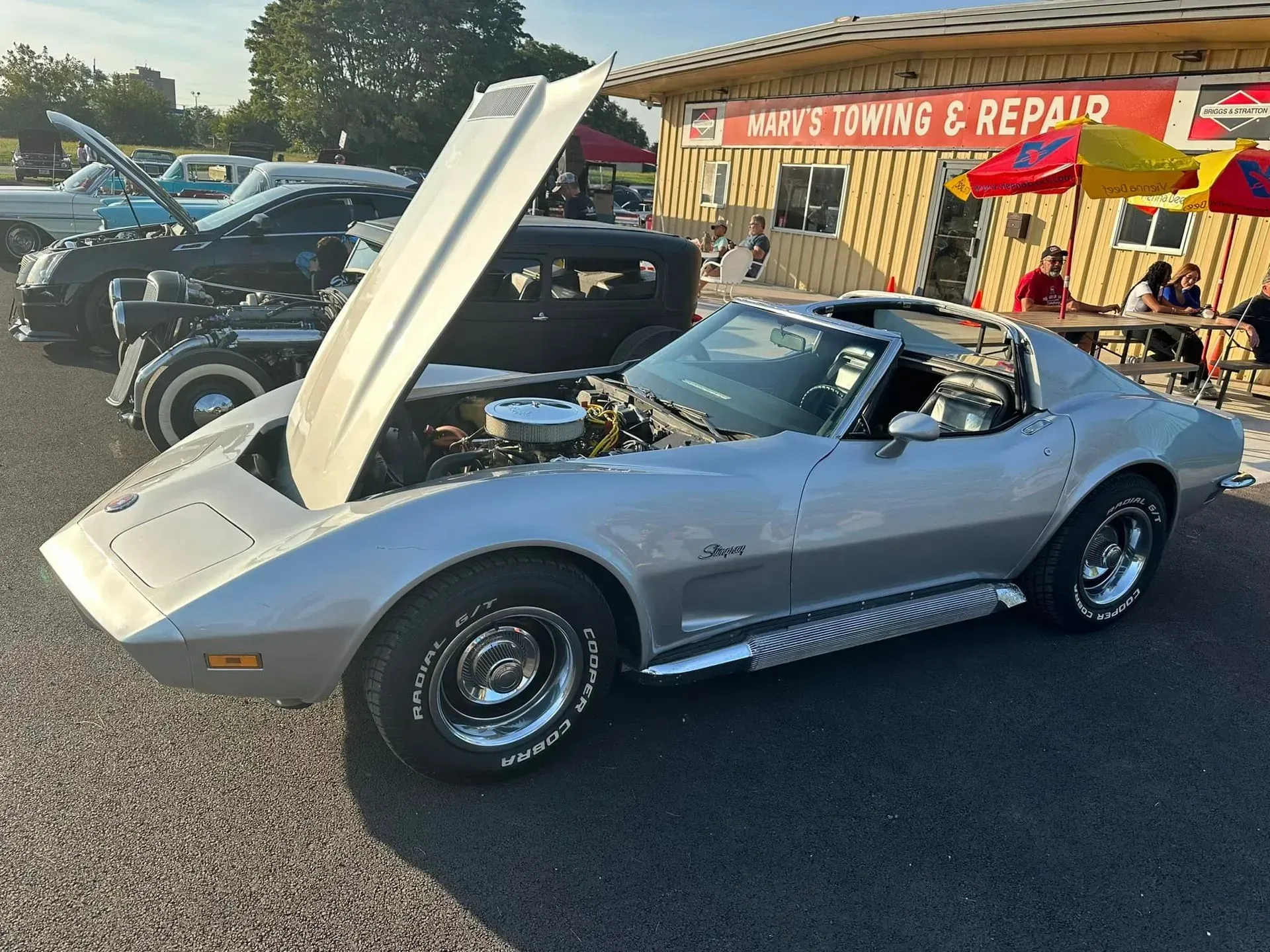 A silver vintage Corvette with its hood open, parked in a lot in front of a towing and repair shop.
