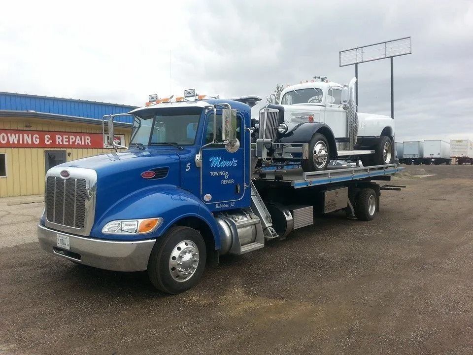 Two blue semi-trucks parked on a gravel lot in front of a building with a red "MARV'S" sign.