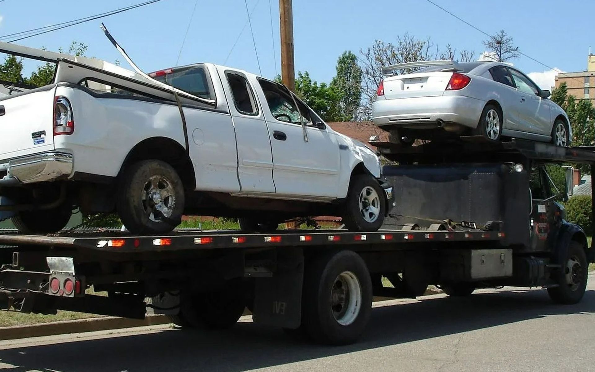 A flatbed tow truck carrying a white pickup truck on the lower level and a silver sedan on the upper rack.