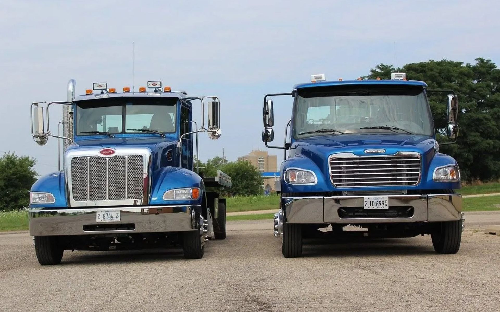 Two blue semi-trucks parked side-by-side on a gravel lot under a bright, cloudy sky.