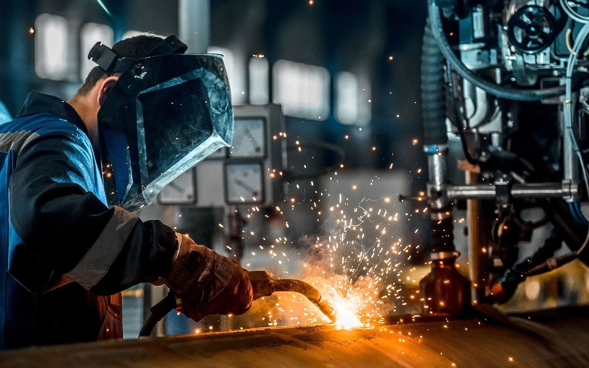 A welder wearing a protective mask works on metal in a factory, creating bright sparks.