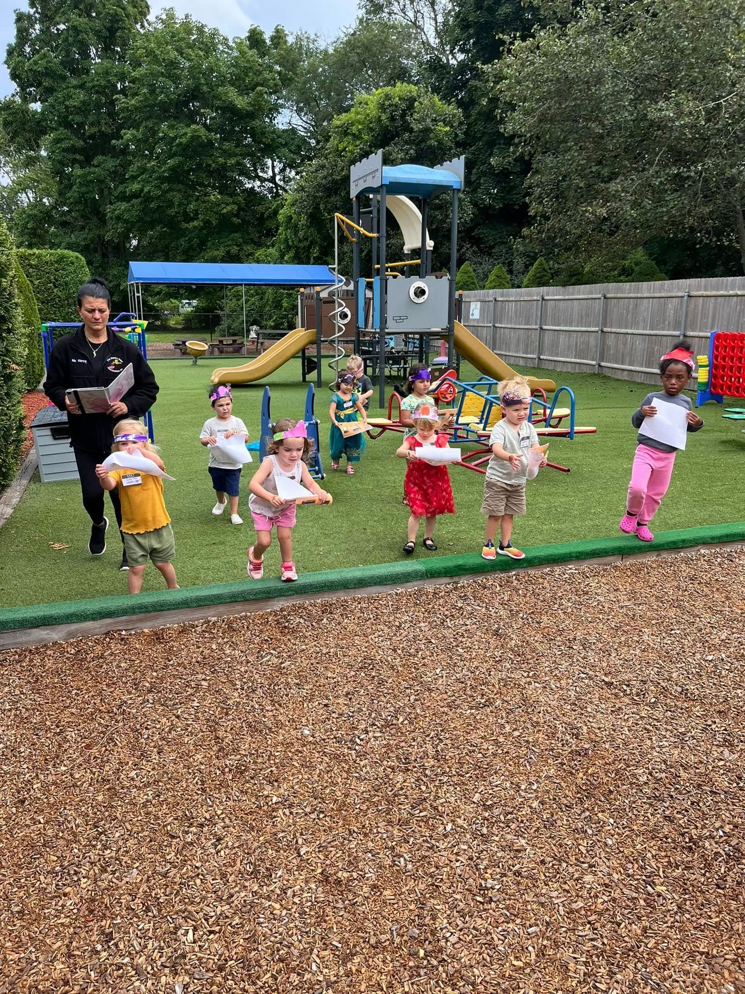 Children holding papers stand in a yard with a teacher, playground equipment in background.