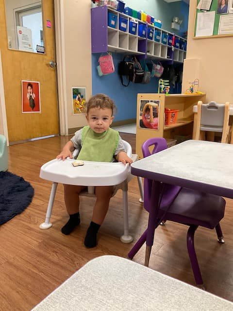 Young child wearing a bib sitting in a high chair at a table in a brightly lit classroom.