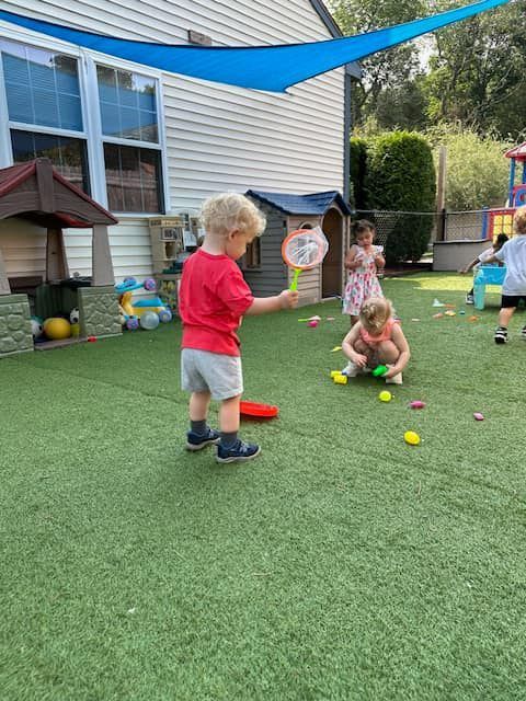Children playing with toys on green turf under a blue shade, near a building.