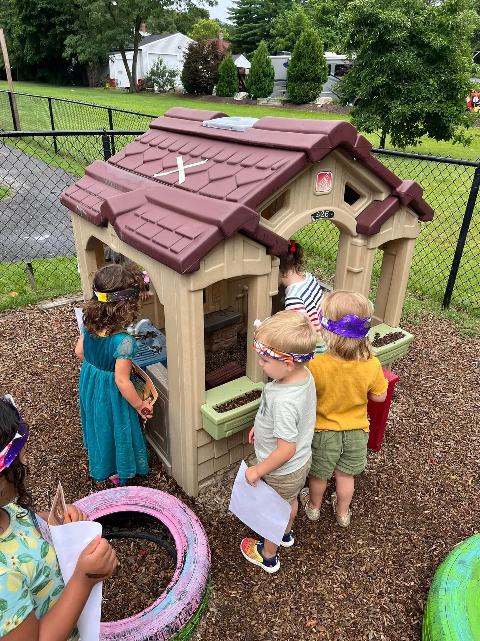 Children playing near a playhouse outdoors. Some wear headbands, holding paper, in a wood chip area.