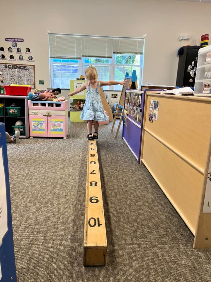 Child balancing on a numbered wooden beam in a classroom, arms outstretched.