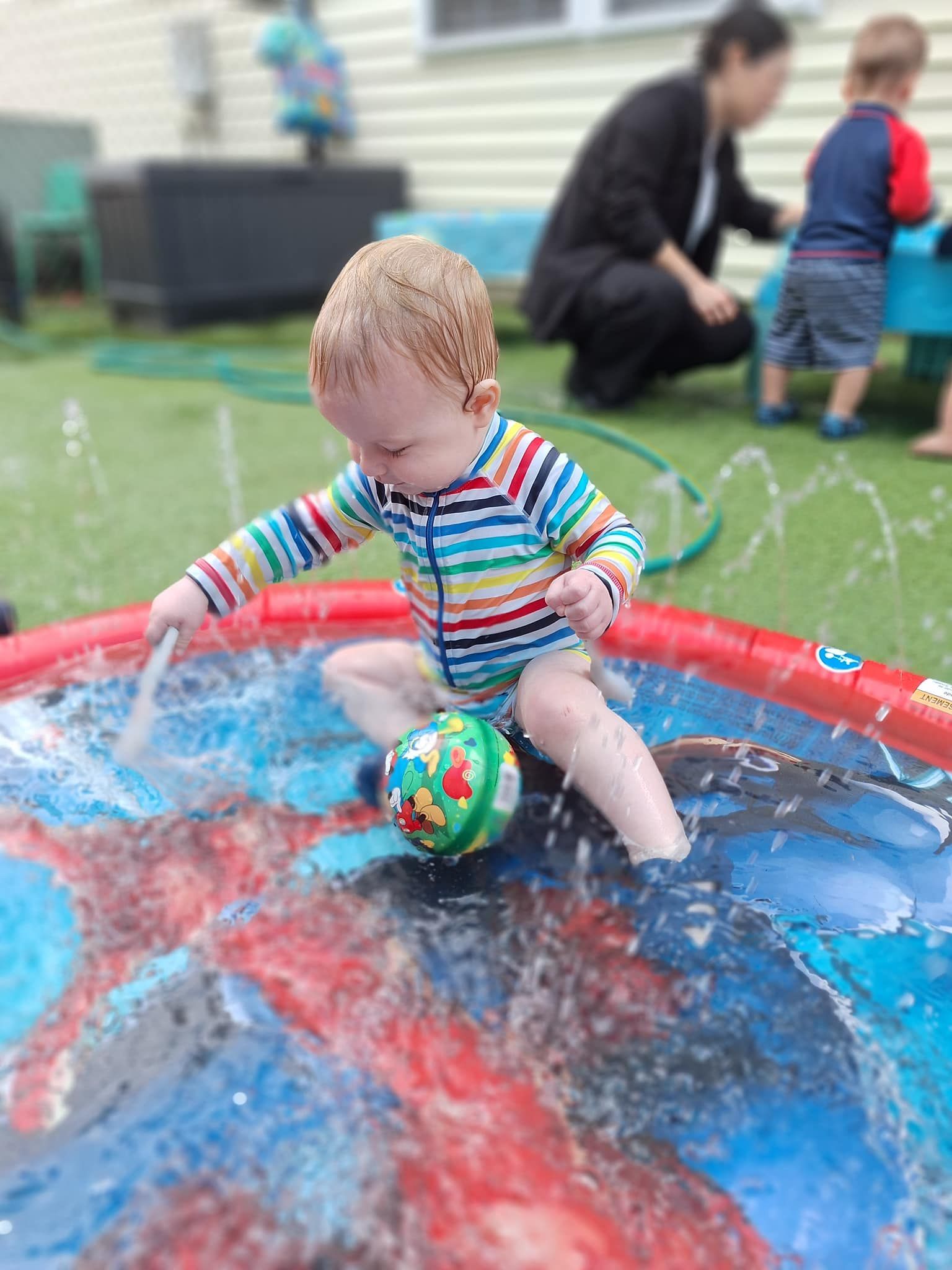 Child in colorful striped swimsuit plays in small pool with a ball, water splashing.