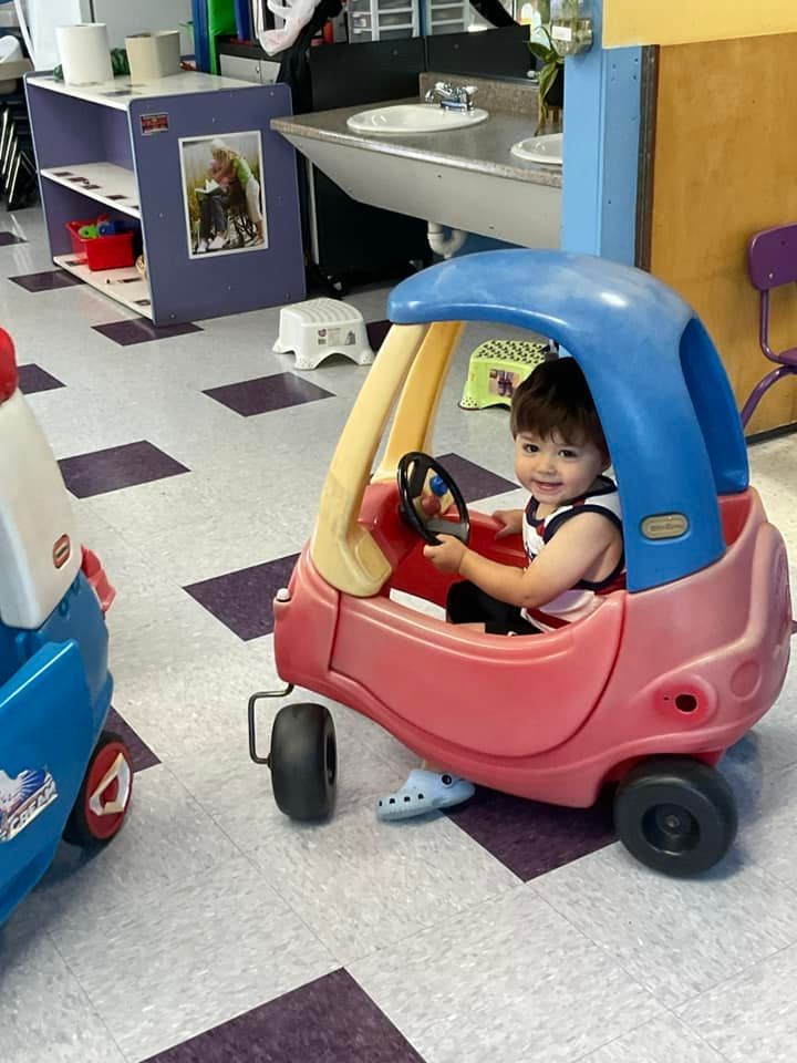 Child in a pink and blue toy car, smiling, in a classroom.