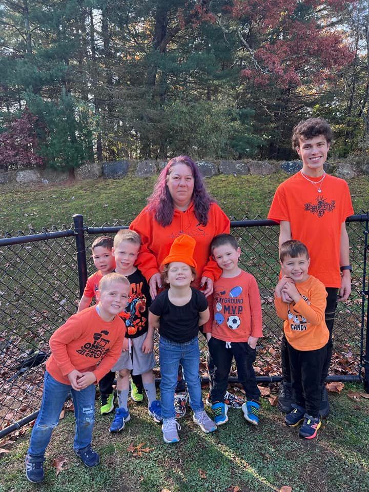 Group of people wearing orange clothing pose near a black fence on a grassy lawn with colorful fall foliage in the background.