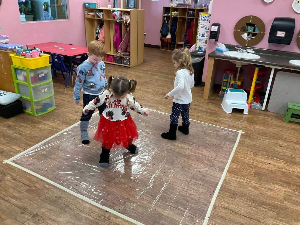 Children playing on a taped square on a wooden floor in a classroom.