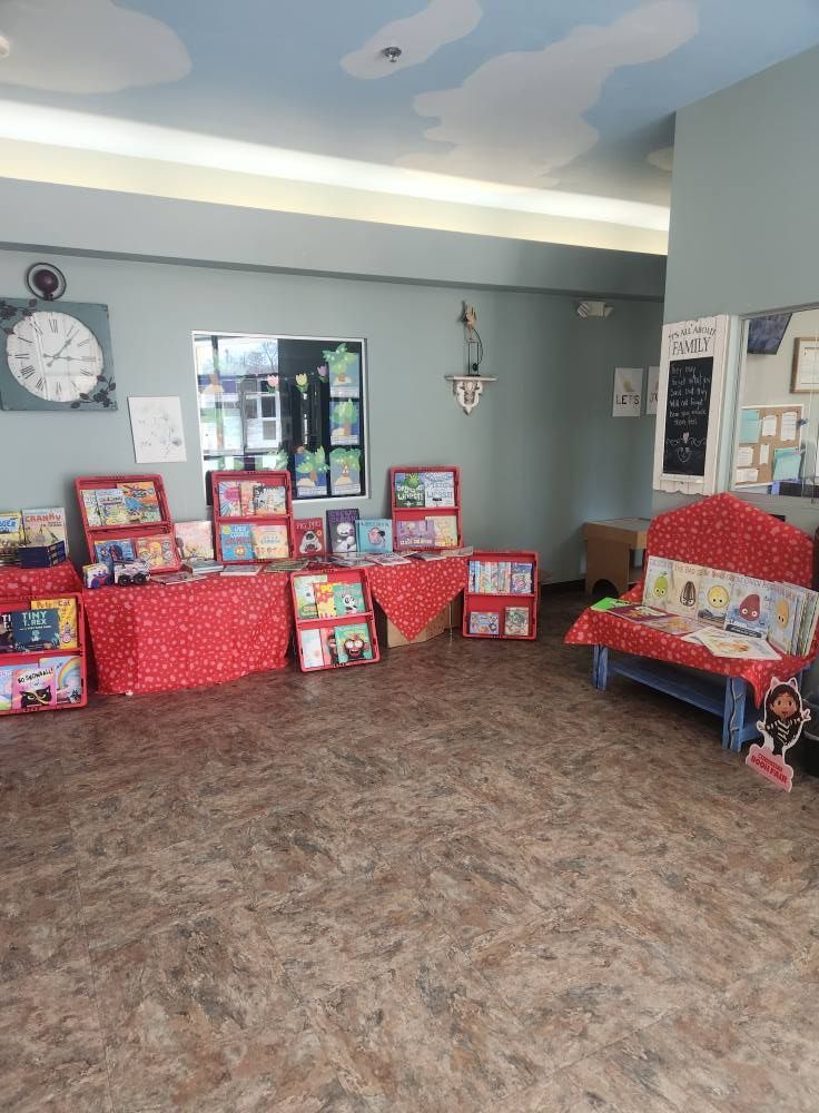 Books displayed on red tablecloths, a reading nook with a bench, and a clock decorate a room.