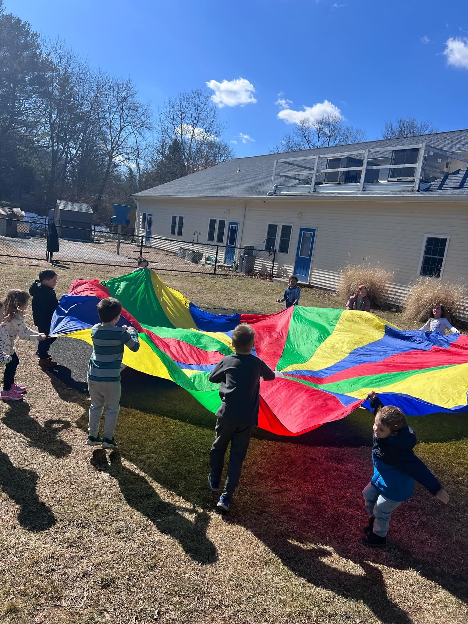 Children playing with a colorful parachute in a grassy area near a building on a sunny day.