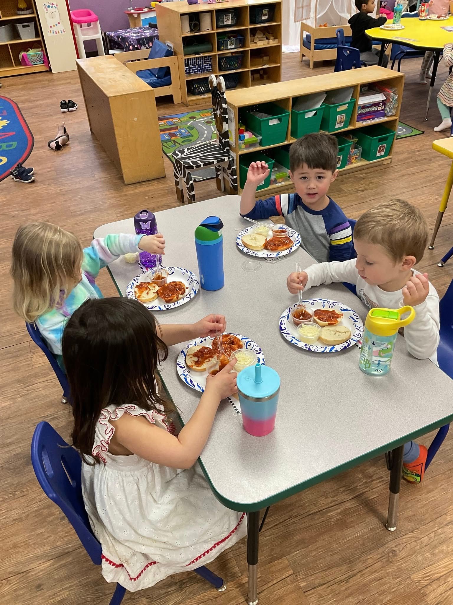 Children eat at a table in a classroom. Plates of food and drinks are on the table.