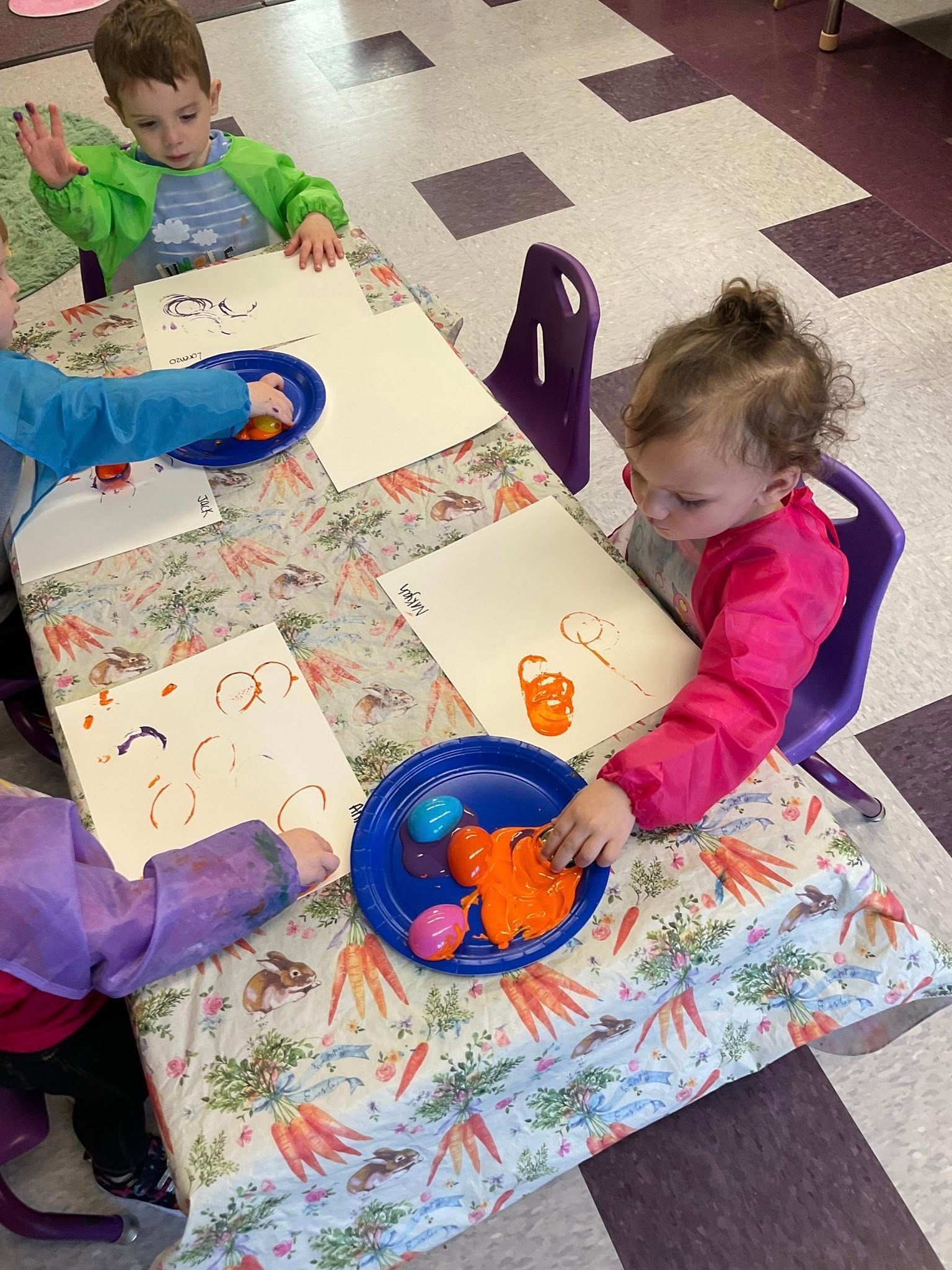 Children painting at a table in a classroom. They are wearing art smocks and using plates of paint.