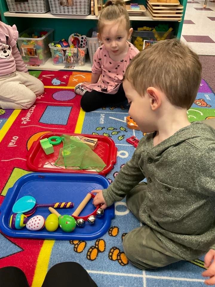 Children playing with toys on a colorful mat. One child reaches for toys in a blue tray; another looks on.