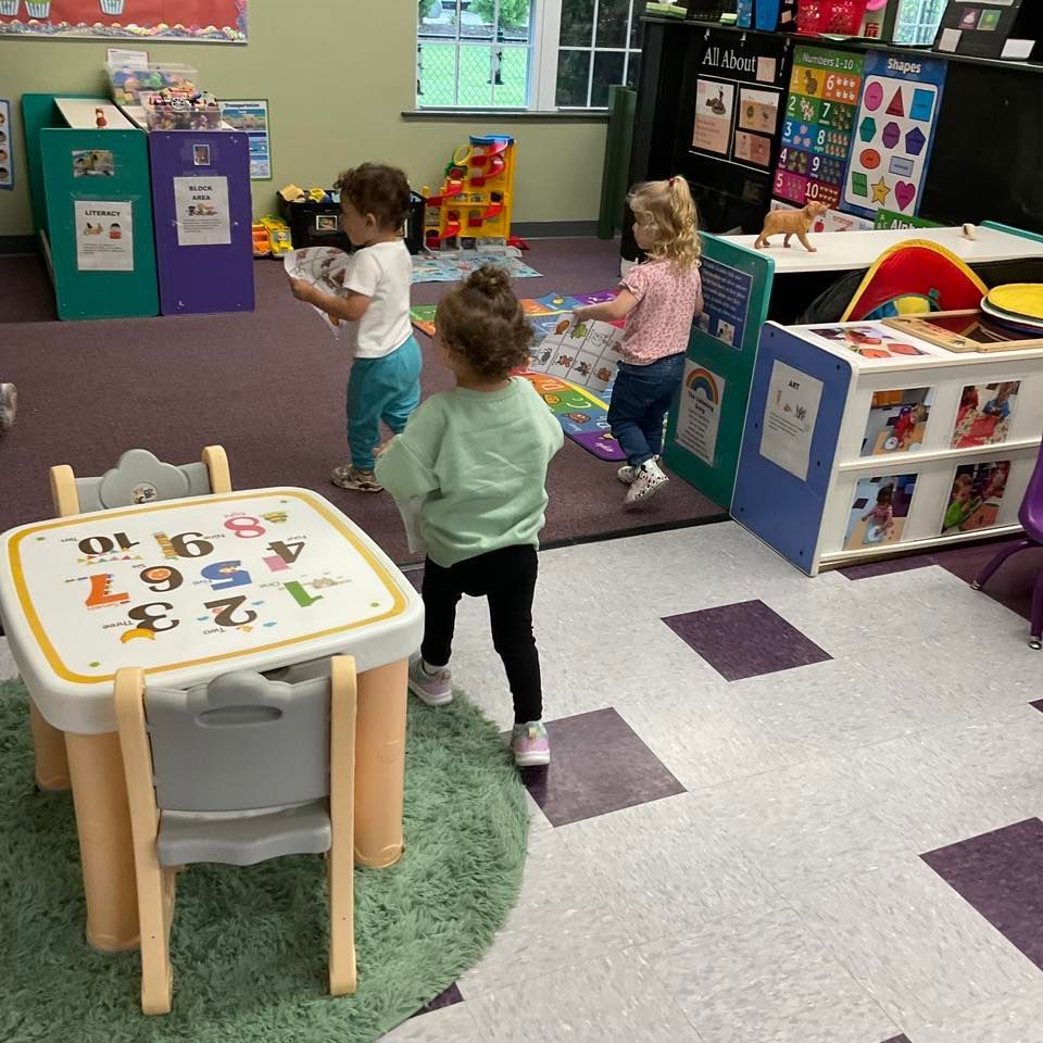Three young children in a brightly colored preschool classroom, playing. A small table is in the foreground.