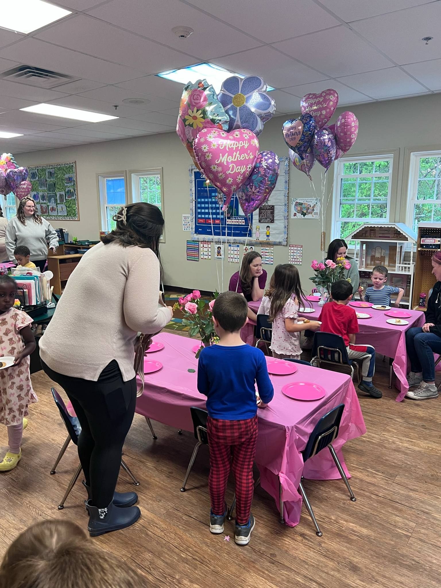 Children at a party with pink tablecloths, balloons, and flowers. A teacher interacts with them.