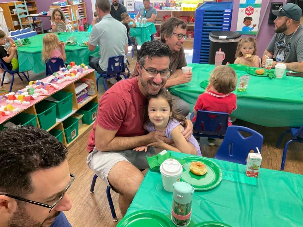 Man and child smile at the camera at a table in a classroom during an event with green tablecloths.