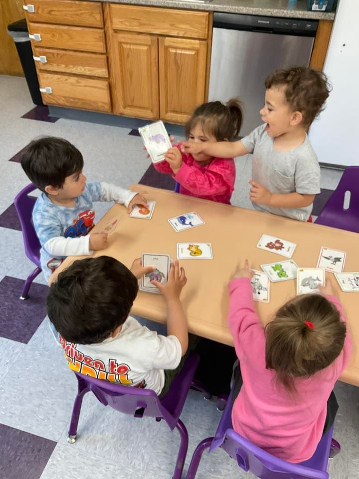 Children at a table playing with flashcards. Purple chairs, wooden cabinets, and a tile floor are visible.