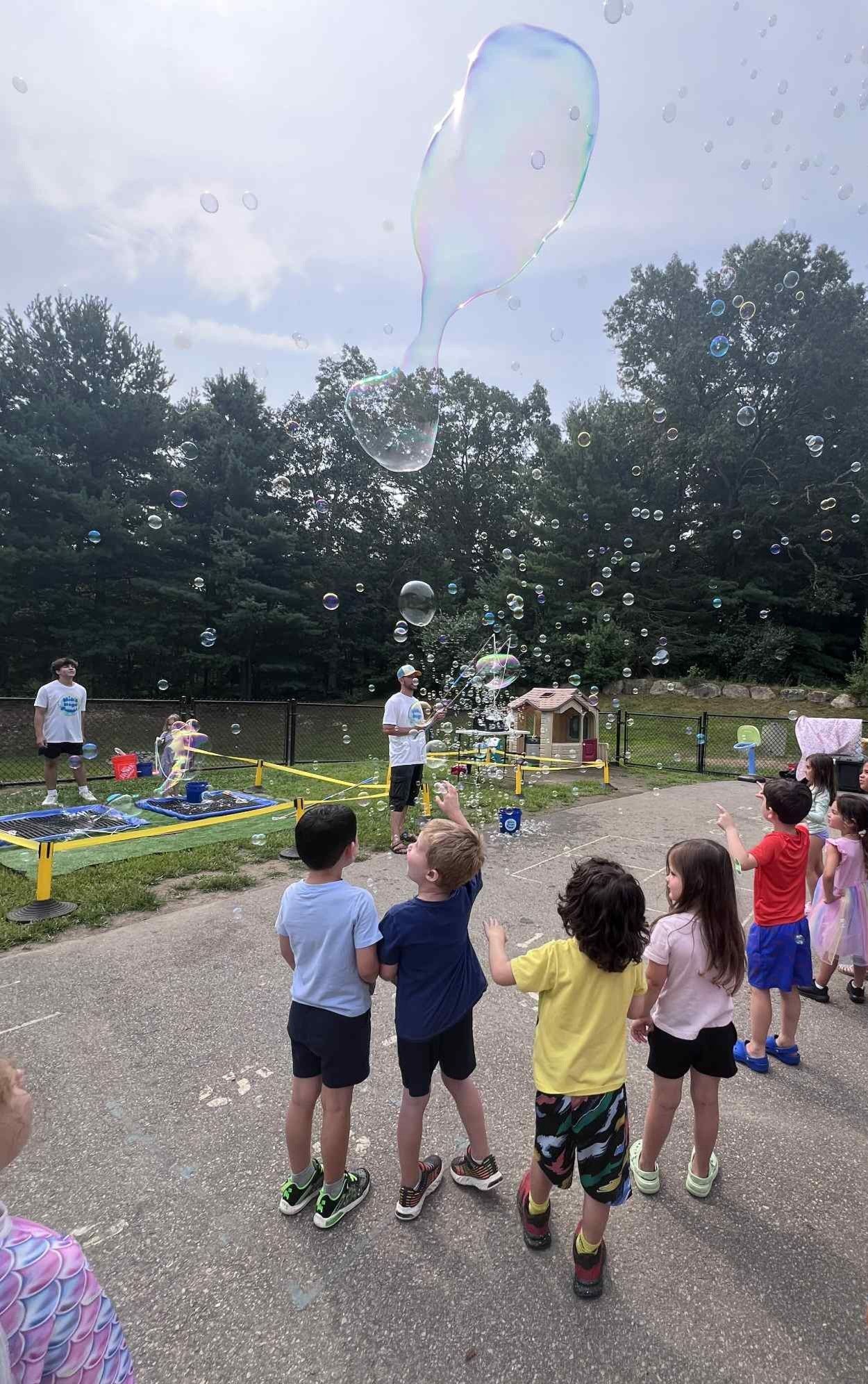 Children watch as large bubbles float into the air; trees and adults are in the background.