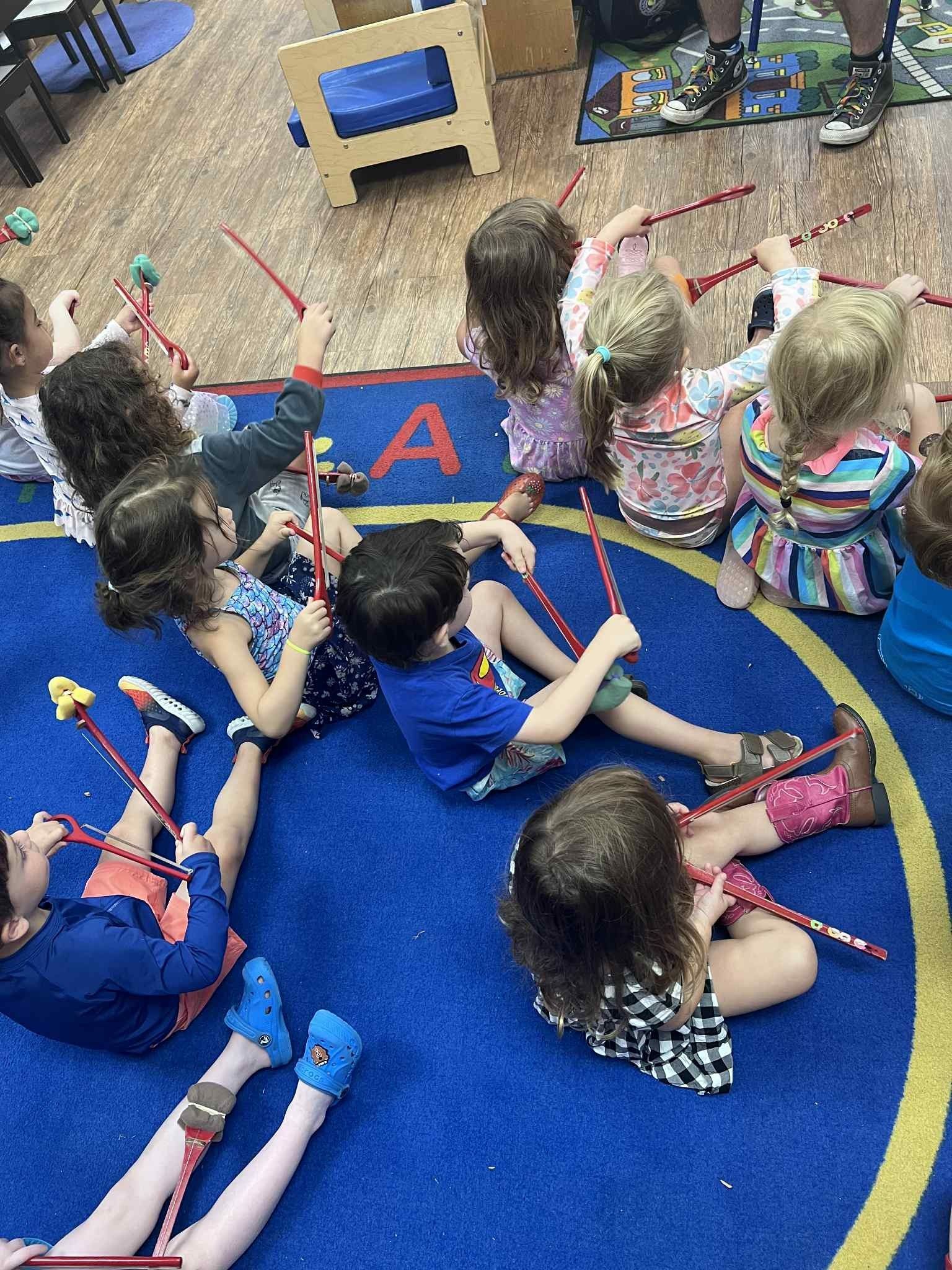 Children sitting in a circle, holding red wands with pom-poms, on a blue carpet.