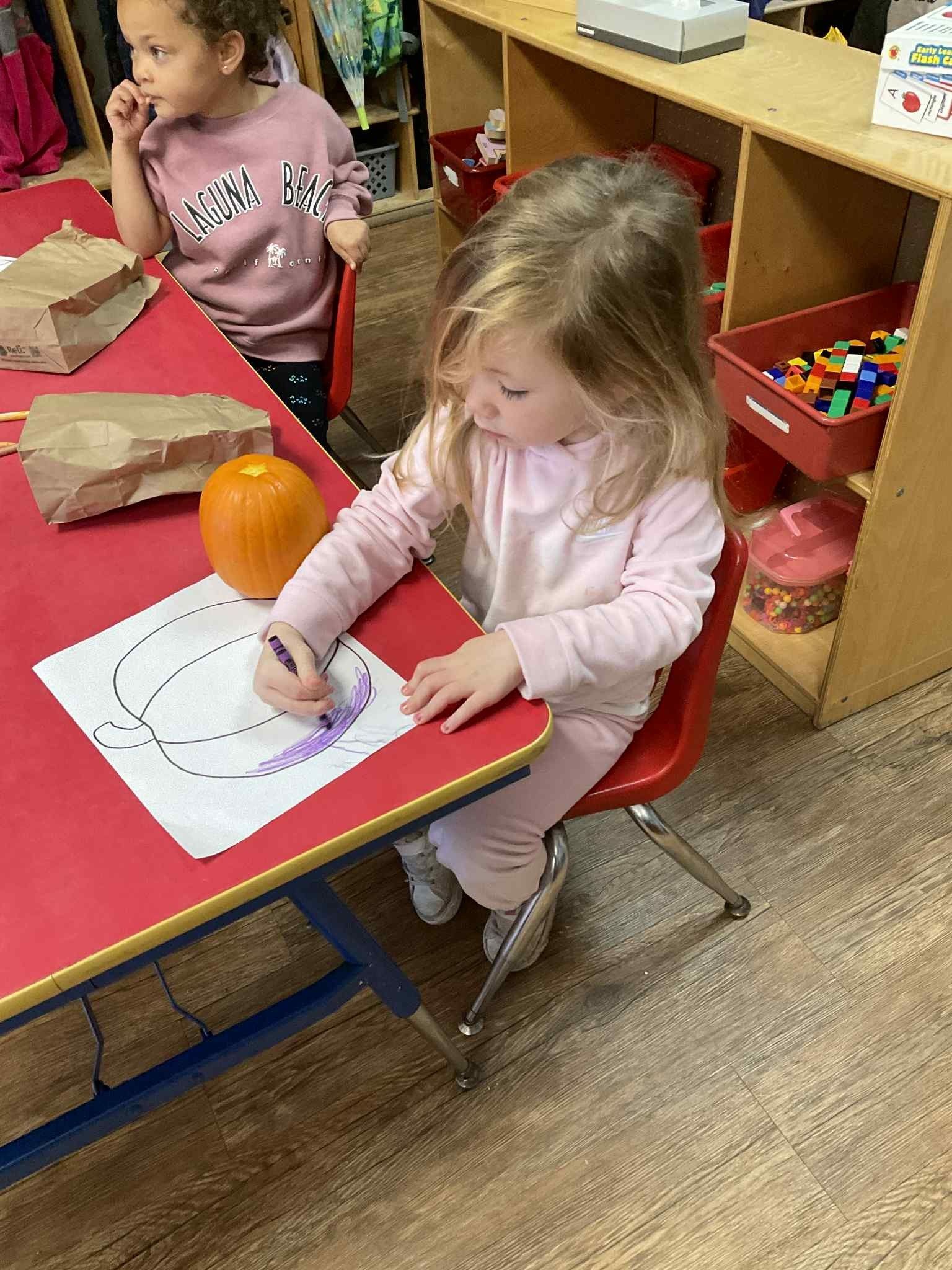 Child seated at a red table, coloring a pumpkin outline.