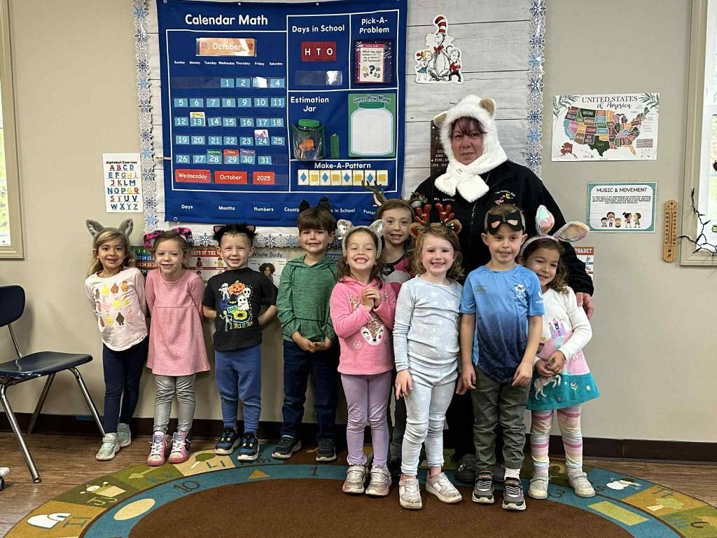 Group of children with teacher, all wearing animal ears, posing in front of a calendar board in a classroom.