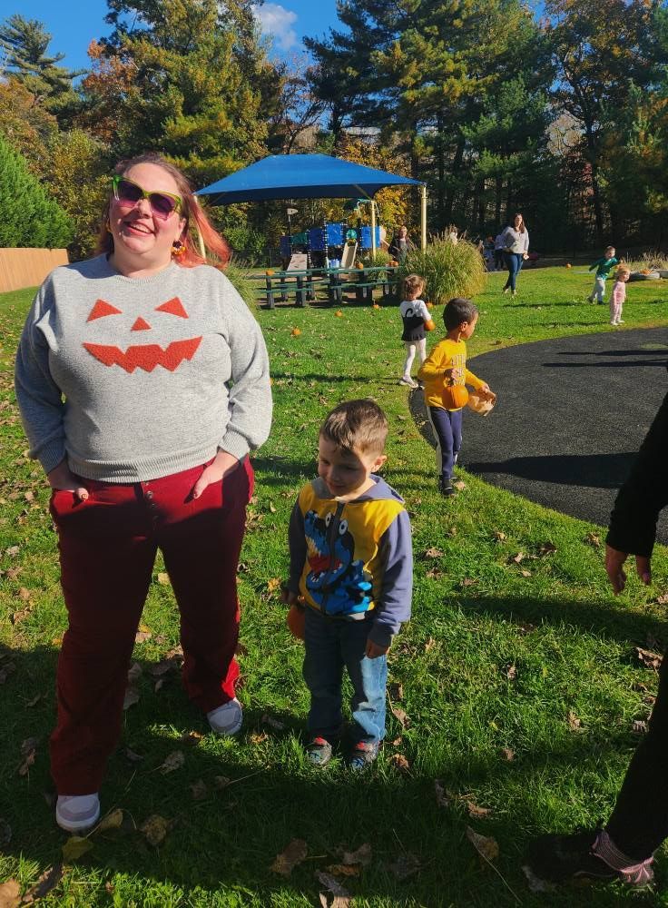 Woman in pumpkin sweatshirt with child, outdoors; other children in background.