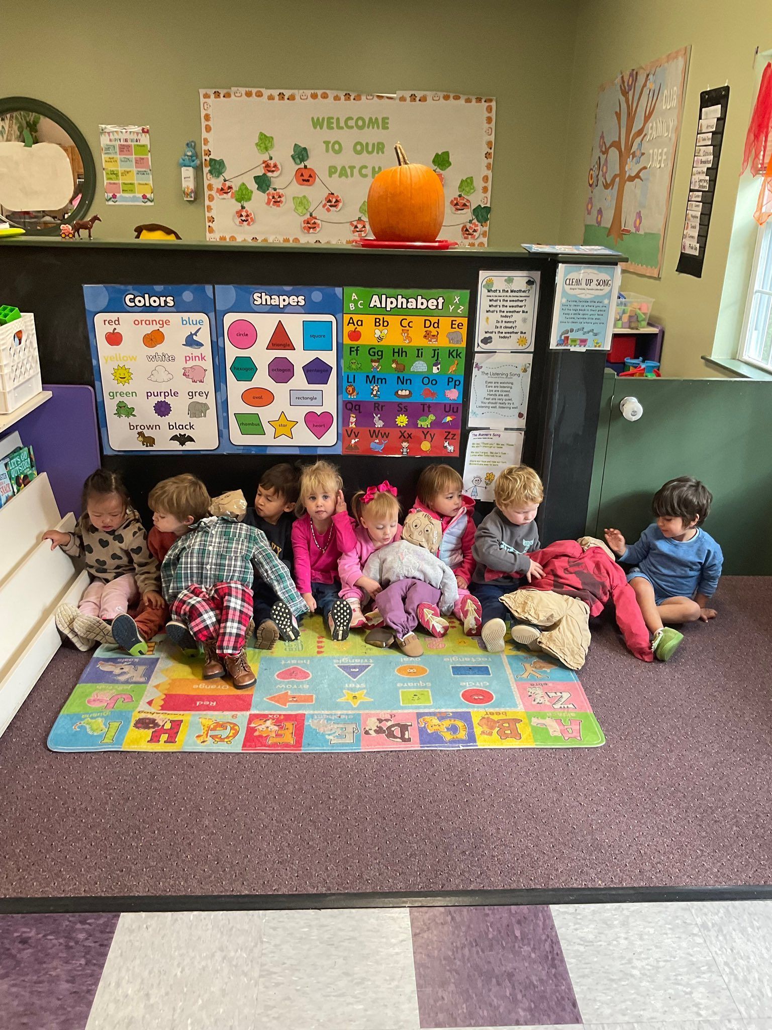 Children seated on mat in a classroom, looking at camera. Pumpkin, charts, and colorful decorations in background.