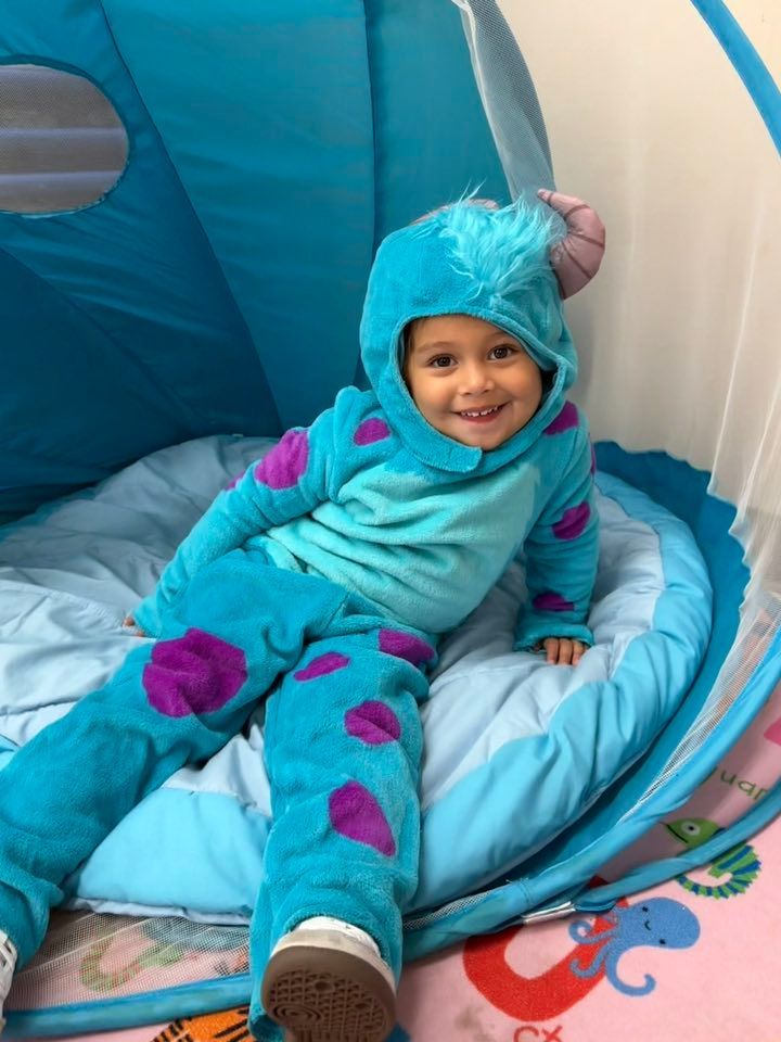 Child wearing a Sully costume, sitting inside a blue tent, smiling.