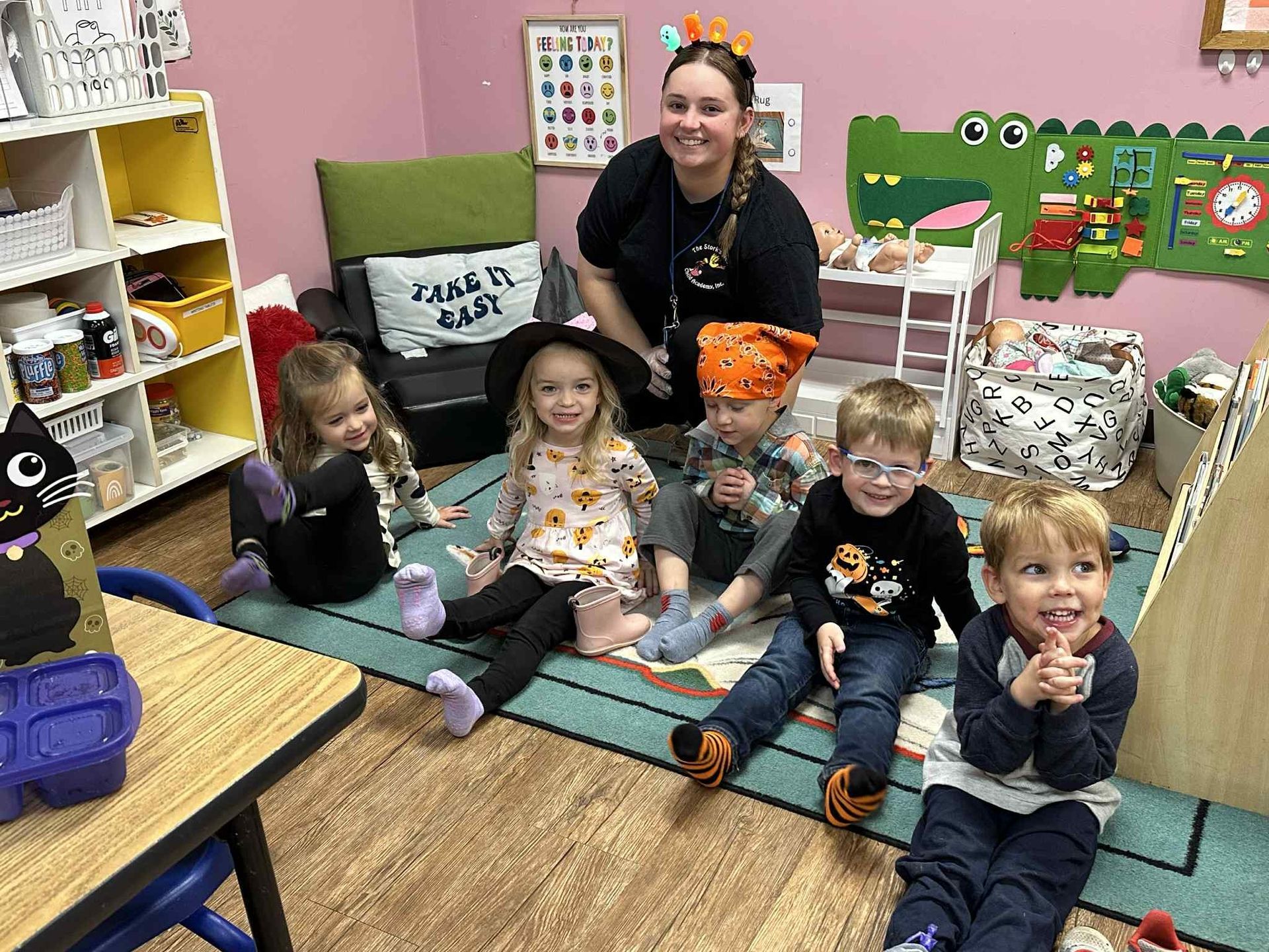 A teacher poses with five children indoors. They are on a carpet. Some wear Halloween costumes.